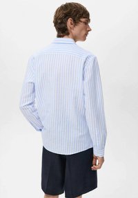 Camicia a righe button-up in azzurro chiaro e rosa, maniche lunghe, colletto standard, tessuto misto cotone e orlo arrotondato. Indossata con pantaloni corti scuri.