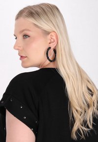 Černé tričko s krátkým rukávem a detaily se cvočky, na modelce s dlouhými blond vlasy a korálkovými černými kruhovými náušnicemi, pohled z boku.