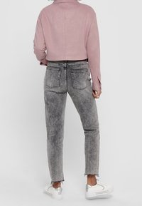 Avkortad rosa mockajacka, grå slitna jeans med fransade fållar och vita sneakers med silverdetaljer. Bakifrån.