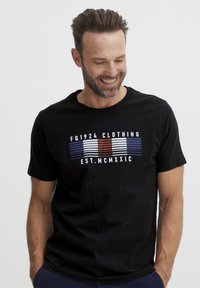 Fekete rövid ujjú póló, amely grafikus nyomtatással rendelkezik, vízszintes piros, fehér és kék csíkokkal, valamint fehér szöveggel. Puha pamut anyagból készült.