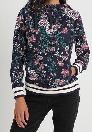 Sweat à capuche bleu marine avec imprimé floral rose et blanc, poignets et ourlet rayés blancs, porté avec un pantalon noir, main dans la poche.