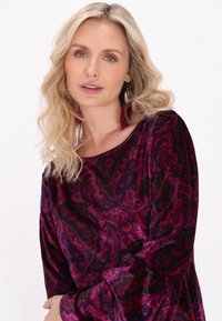 Abito in velluto viola con motivo paisley, scollatura a barca e maniche lunghe. Il modello indossa orecchini di piume rosse. Texture liscia.