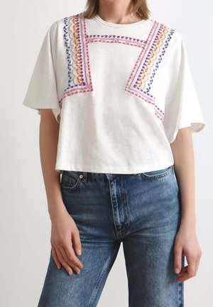 Femme portant un haut blanc à manches courtes avec des motifs géométriques colorés brodés, associé à un jean en denim bleu taille haute.