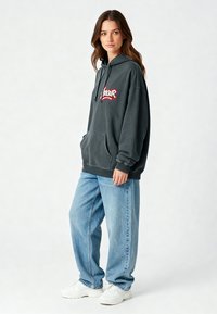 Felpa oversize grigio carbone con tasca frontale e logo ricamato "Amour" in rosso. Abbinata a jeans larghi blu chiaro e sneakers bianche.