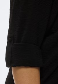 Μαύρο πουκάμισο με υφή στην επιφάνεια, με μανσέτες ριγμένες και λεπτομέρεια με κουμπιά. Το ύφασμα φαίνεται ελαφρύ και ελαφρώς ζαρωμένο.