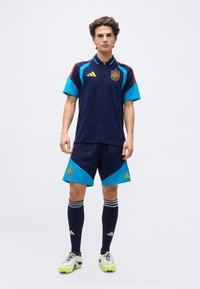 Young man stands wearing navy and blue soccer kit with Spain crest, striped navy socks, and cleats against plain white background.
