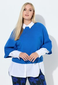 Blauer Strickpullover mit kurzen Ärmeln, getragen über einem weißen Hemd mit Kragen, kombiniert mit gemusterten marineblauen Hosen mit Blumenmotiven.