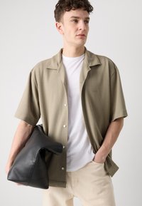 Rövidujjú khaki ing, texturált anyagból, gombos záródással és kis logóval. A modellen fekete bőr táska van, fehér pólót visel.