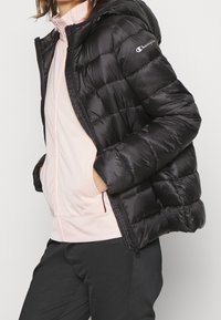 Person som bär en svart Champion puffjacka över en ljusrosa dragkedjetröja med händerna i jackfickorna, mot en enkel bakgrund.