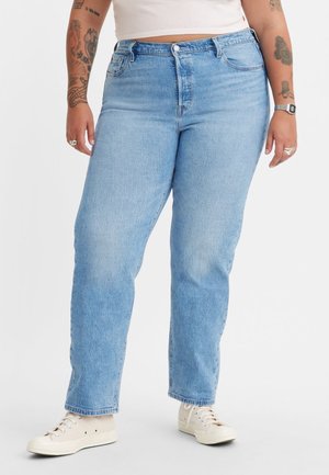 Kurvige Person, die hellblaue High-Waist-Jeans, weiße Sneakers, ein weißes bauchfreies Oberteil sowie sichtbare Arm-Tattoos und Accessoires trägt.