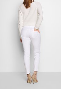 Bílé přiléhavé džíny s hladkou texturou a našitými zadními kapsami, doplněné světle zbarveným svetrem s dlouhými rukávy a béžovými podpatky.
