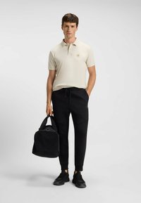 Beige polo-shirt met een logo, zwarte joggerbroeken en zwarte sportschoenen. Houdt een zwarte tas vast met een afgeronde bodem en dubbele hengsels.