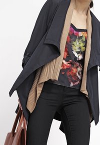 Cardigan marron à côtes superposé sur un haut à imprimé floral, veste bleu marine drapée et pantalon noir. Sac fourre-tout en cuir marron. Textures lisses.