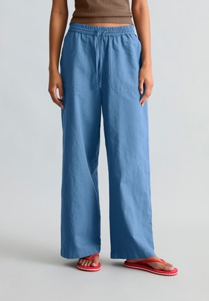 Osoba nosząca luźne niebieskie spodnie ze ściągaczem w talii i brązowy bezrękawnik, stojąca na szarej podłodze w czerwonych klapkach.