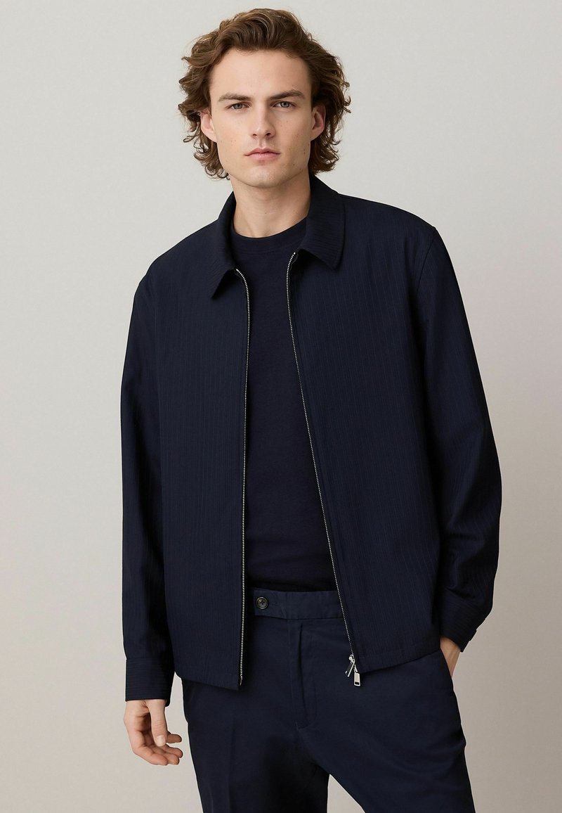 Chaqueta de felpa azul marino con cremallera, cuello de camisa y rayas verticales. Textura suave con mangas largas, llevada sobre una blusa y pantalones a juego en azul marino.