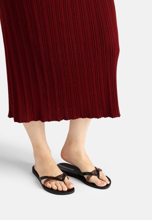 Füße, die schwarze und braune gemusterte Flip-Flop-Sandalen tragen, mit einem gerippten dunkelroten Rock, der von der Mitte der Wade abwärts sichtbar ist.