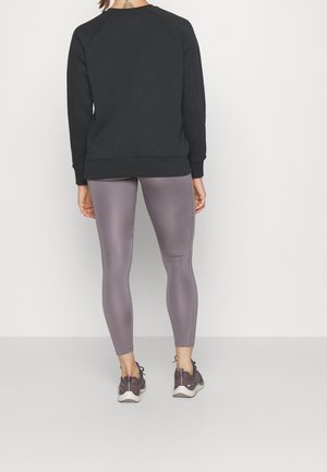 Person steht mit dem Rücken zur Kamera, trägt ein schwarzes langärmeliges Oberteil, graue enganliegende Leggings und dunkle Sportschuhe vor einem einfarbigen hellen Hintergrund.