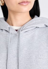 Gros plan d'une personne portant un sweat à capuche gris clair avec le logo "cK" sur la poitrine et des boucles d'oreilles créoles dorées, montrant les lèvres et le cou.