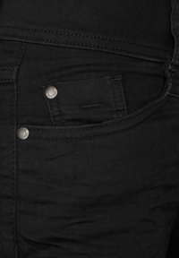 Jeans de mezclilla negra con una textura suave, que presentan herrajes plateados, costuras en los bolsillos frontales y un sutil detalle descolorido en la tela.