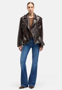 Brązowa skórzana kurtka motocyklowa z klapowymi kieszeniami, szerokim kołnierzem i naramiennikami, zestawiona z niebieskimi rozszerzanymi dżinsami i brązowymi butami.