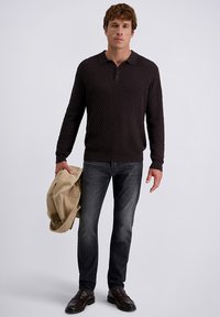 Mann in einem schwarzen, strukturierten Langarm-Polo, dunklen grauen Jeans und schwarzen Schuhen, der eine beige Jacke vor einem schlichten weißen Hintergrund hält.