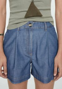 Pantalones cortos de mezclilla con un corte relajado, color azul claro, pliegues frontales y costuras visibles. Combinados con una camiseta sin mangas de canalé verde.