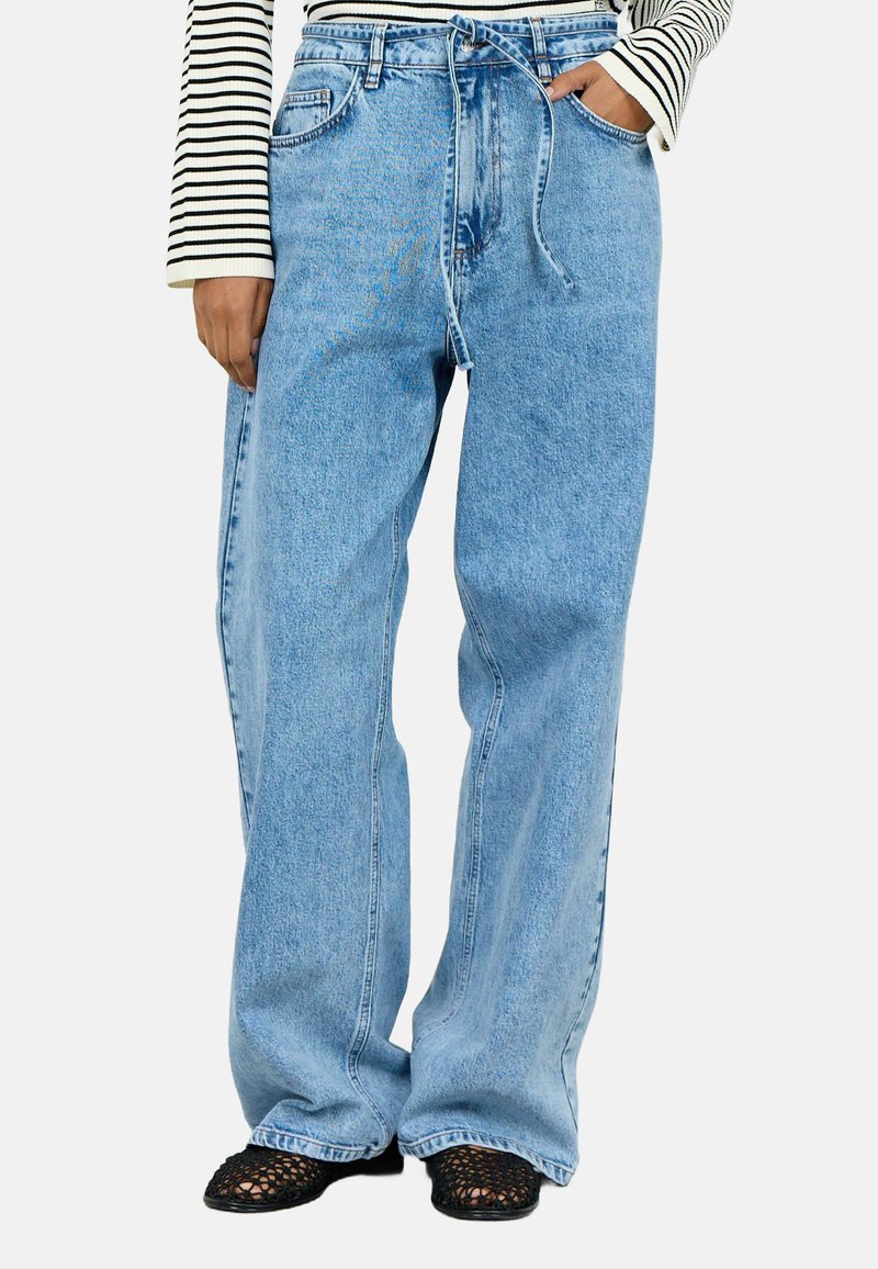 Helle blaue weite Denim-Jeans mit Kordelzugbund, frontalen Taschen und entspannter Passform, kombiniert mit schwarzen Mesh-Schuhen.