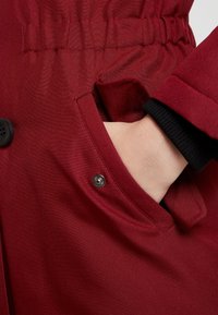 Röd kappa med texturerad tyg, som har en ficka med knäppning och elastisk midja. Inkluderar svarta manschettdetaljer och tryckknappsstängning.