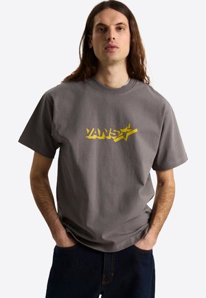 Joven con pelo largo que lleva una camiseta gris de Vans con un logo amarillo, manos en los bolsillos, sobre un fondo claro y liso.