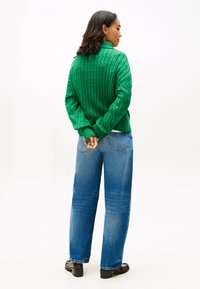 Zöld bordázott kötött pulóver magas gallérral, magas derekú, kék egyenes szárú farmerrel és sötét loaferekkel párosítva, hátulról nézve.