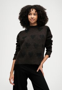 Καφέ πλεκτό πουλόβερ με μαύρα σχέδια καρδιάς, στρογγυλή λαιμόκοψη και τρία τέταρτα μανίκια. Μαλακό, υφή ύφασμα, συνδυασμένο με μαύρα παν trousers.