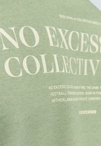 Grön bomulls-t-shirt med vit text. Texten inkluderar "NO EXCESS" och andra märkesdetaljer i varierande teckenstorlekar, slät textur, avslappnad design.