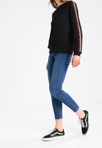 Czarna bluza z kolorowymi paskami na rękawach, w połączeniu z niebieskimi obcisłymi dżinsami i czarnymi trampkami Vans. Casulowy strój o minimalistycznym wzornictwie.