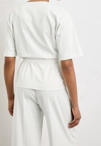 Top bianco a maniche corte con un fit morbido e vita annodata. Tessuto liscio e opaco. Dettaglio posteriore presenta un collo alto e senza decorazioni.