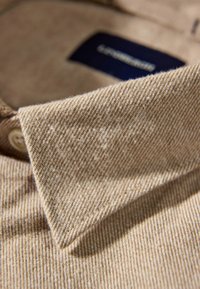 Beige overhemd met kraag en een gestructureerde stof. De kraag is scherp afgebiesd en een knoop is zichtbaar nabij de kraag.