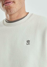 Zbliżenie osoby w białej polarowej bluzie z małą czarną koroną i logo "QS" na lewej piersi.