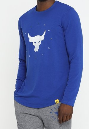 Man draagt een blauw shirt met lange mouwen met een witte stierschedel en een cirkelvormige tekst "ALL DAY HUSTLE", gecombineerd met grijze joggingbroek.