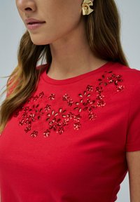 Camiseta de algodón roja con diseño de corte floral adornado con lentejuelas rojas en el escote. Mangas cortas y cuello redondo.
