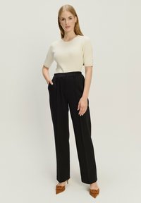 Hellbeige gerippte Strickoberteil mit kurzen Ärmeln, kombiniert mit hoch taillierten schwarzen Faltenhosen und braunen spitzen Absätzen.