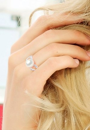 Bague en argent avec une perle blanche lisse au centre, rehaussée de détails scintillants et d'un design délicat. Main reposant doucement sur les cheveux.