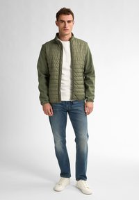 Giacca trapuntata verde oliva con colletto a camicia, indossata sopra un maglione bianco, abbinata a jeans azzurri chiari e sneakers bianche.