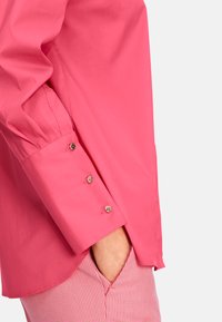 Fuchsia cotton shirt with long sleeves and buttoned cuffs, featuring three decorative buttons and an angular hem. Paired with pink striped pants.