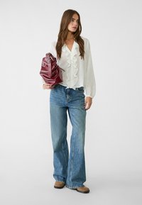 Vit rynkad blus, ljusblå vida jeans och en bordeauxfärgad läderväska med spännhandtag. Modellen står med avslappnade händer.