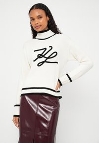 Beli pleteni pulover z visokim ovratnikom, na katerem je izrazit črn logotip in kontrastni črni trakovi na rokavih in spodnjem robu. Pod njim sijoča bordo krilo.