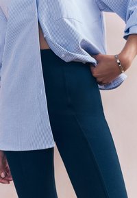 Modro-bíle pruhovaná košile částečně zastrčená do tmavě modrých slim fit legín. Stříbrný náramek na zápěstí. Hladká látka a elegantní design.