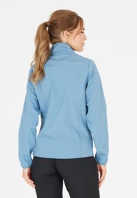 Veste softshell bleu clair avec un col haut, des manches longues et une coupe ajustée. Texture lisse, poignets élastiques et détails de coutures.