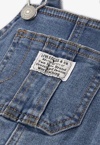 Τζιν φορμάκι με ξεθωριασμένο μπλε χρώμα, ορατή ραφή και ορθογώνια υφασμάτινη ετικέτα που αναγράφει το εμπορικό σήμα και τις πληροφορίες κατασκευής.