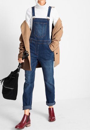 Salopette in denim blu scuro con bretelle regolabili, tasche frontali e risvolti; abbinata a un dolcevita bianco e un cappotto marrone. Stivaletti rossi alla caviglia.