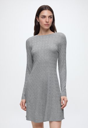 Mujer con cabello largo y castaño, vestida con un vestido ajustado de manga larga de punto trenzado en gris claro, de pie delante de un fondo blanco liso.