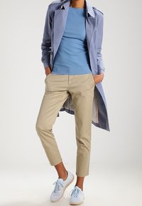 Person som bär en ljusblå tröja, lavendelfärgad trenchcoat, beige kapade byxor och vita sneakers med ljusblå snören, med händerna i fickorna.